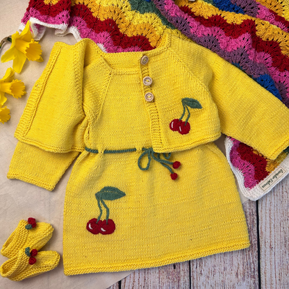 Yellow knitted baby outfit with cherry details on a wooden surface