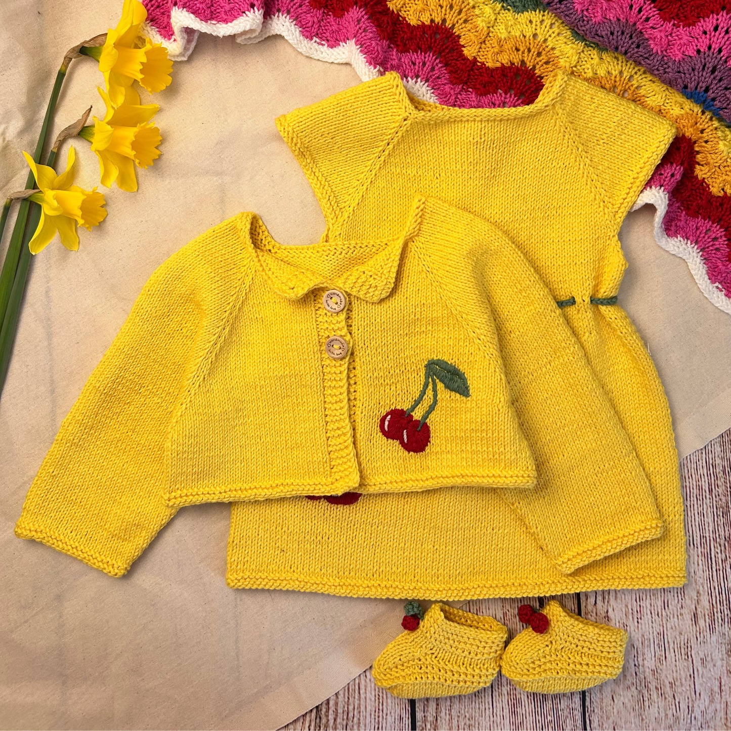 Yellow knitted baby outfit with cherry design on a textured surface with flowers.