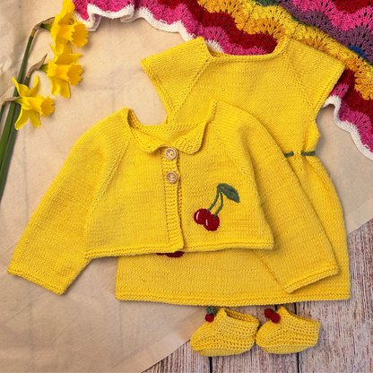 Yellow knitted baby outfit with cherry design on a textured surface with flowers.