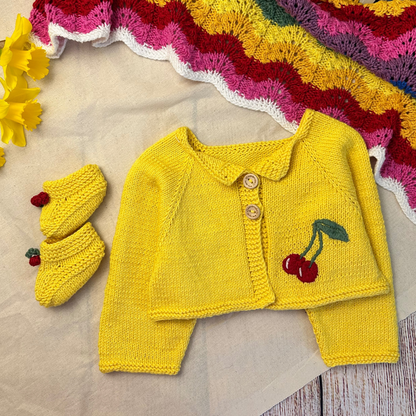 Yellow knitted baby outfit with cherry details on a textured surface.