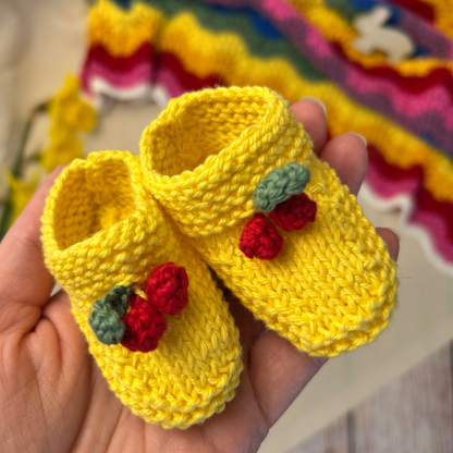 Yellow knitted baby shoes with red berries held in a hand, colorful background