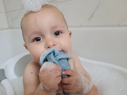 Baby in a bathtub holding a blue towel with soap suds.