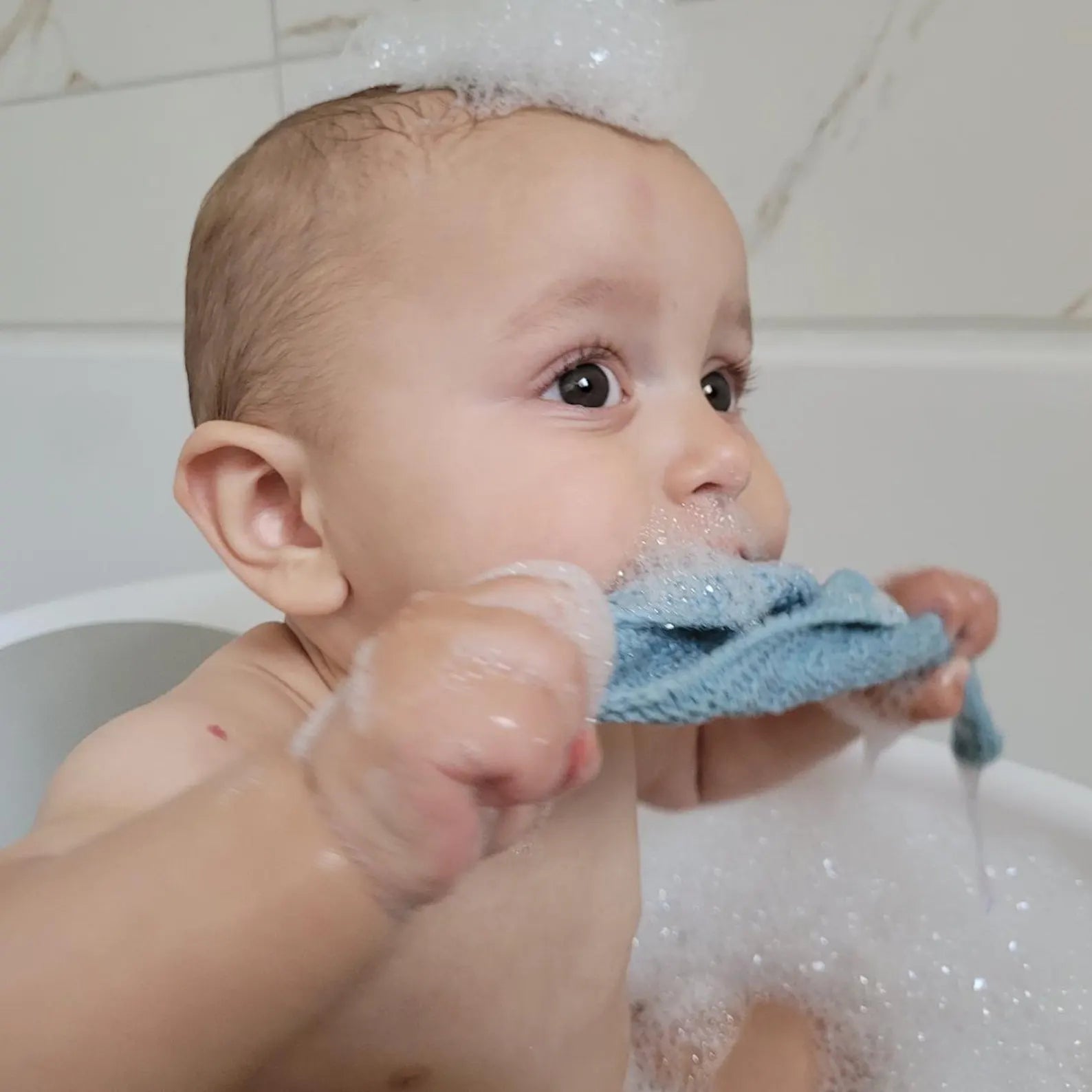 Baby in a bathtub with bubbles holding a blue towel