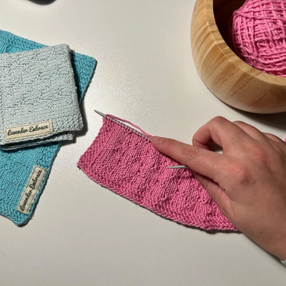 Hand knitting a pink fabric with a wooden bowl of yarn and two knitted items labeled 'Lavender Enchante' on a white surface.