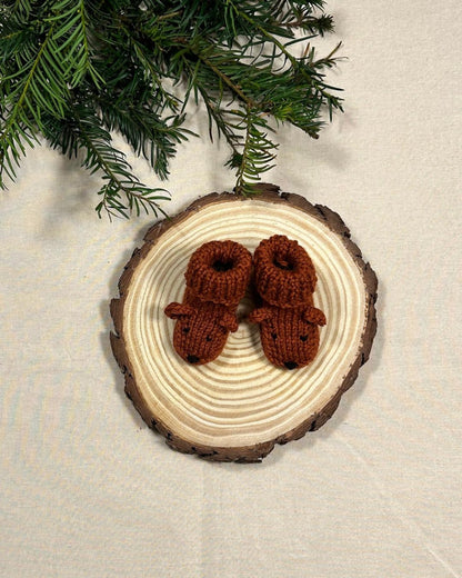 Pair of brown knitted mitten ornaments on a wooden tree slice with greenery in the background.