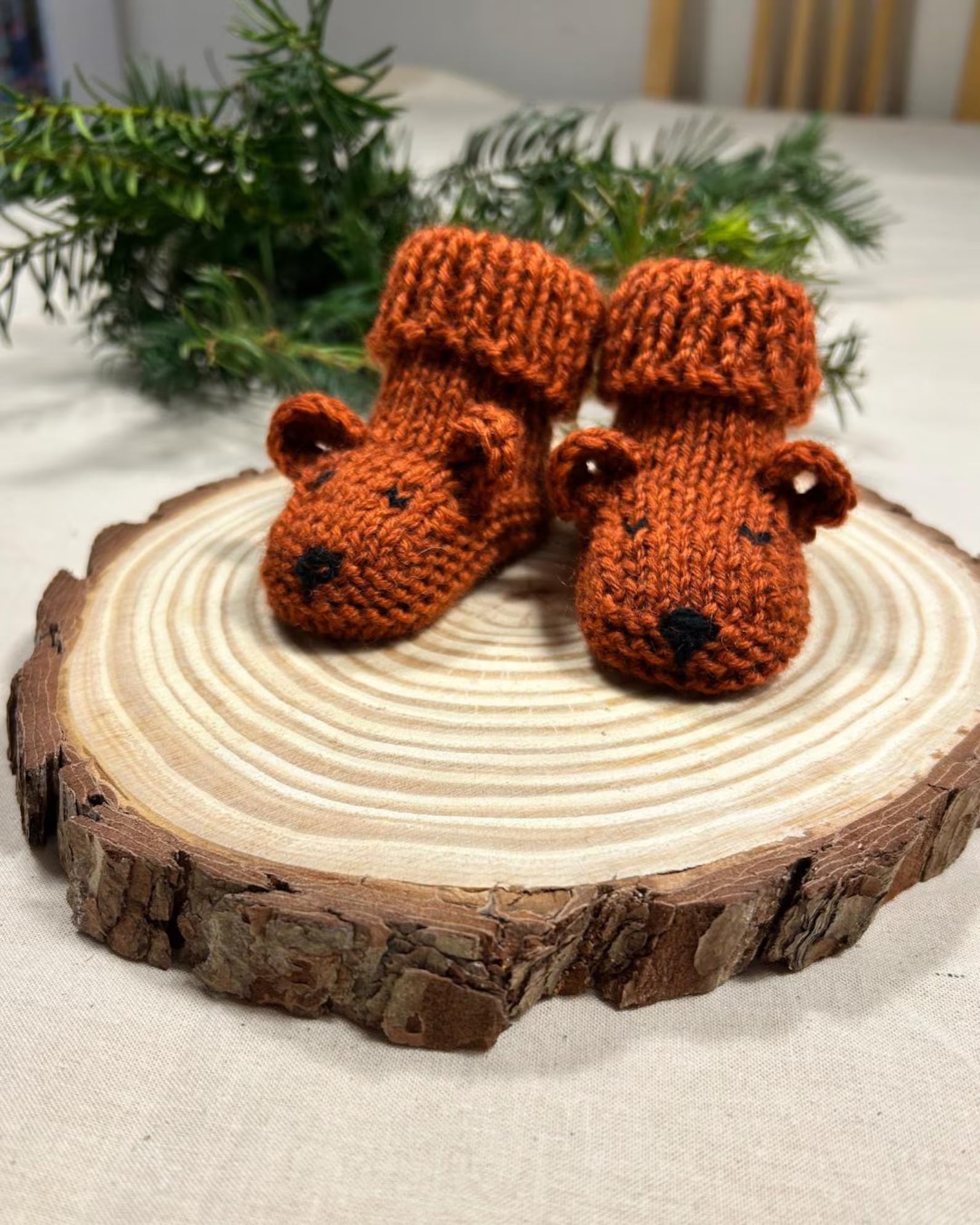 Pair of knitted brown bear booties on a wooden log with greenery in the background