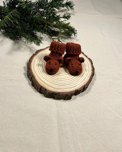 Knitted reindeer boots on a wooden disc with a Christmas tree branch in the background.